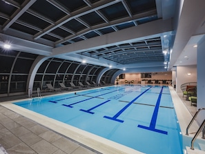 An indoor swimming pool with clear lane markings, a high ceiling with skylights, and lounge chairs along the poolside.