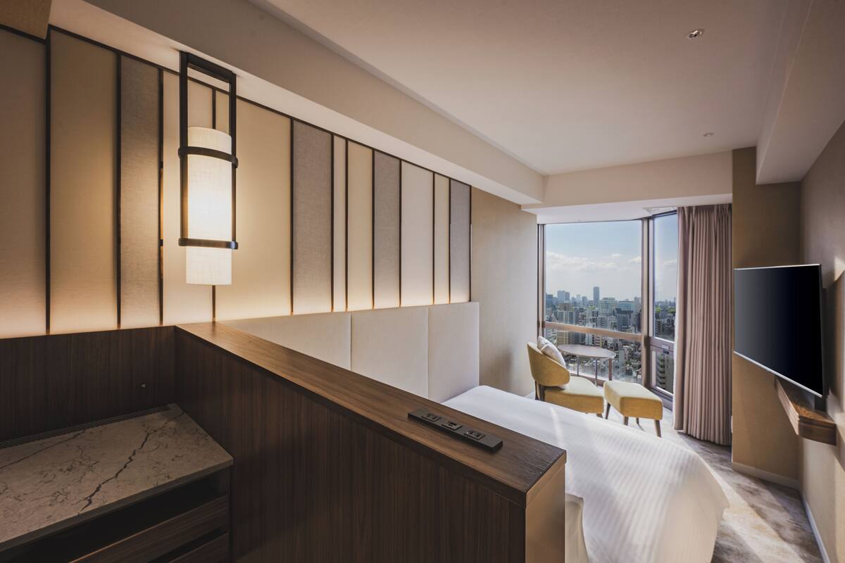 A hotel room with a bed, a desk, a TV, a city view, and a large window.
