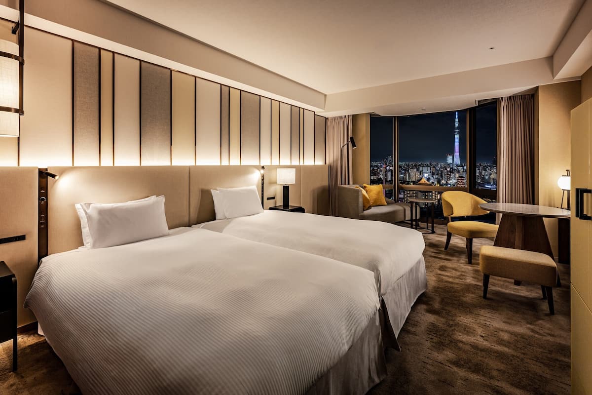 A hotel room with two beds, a desk, a chair, and a view of a cityscape at night.