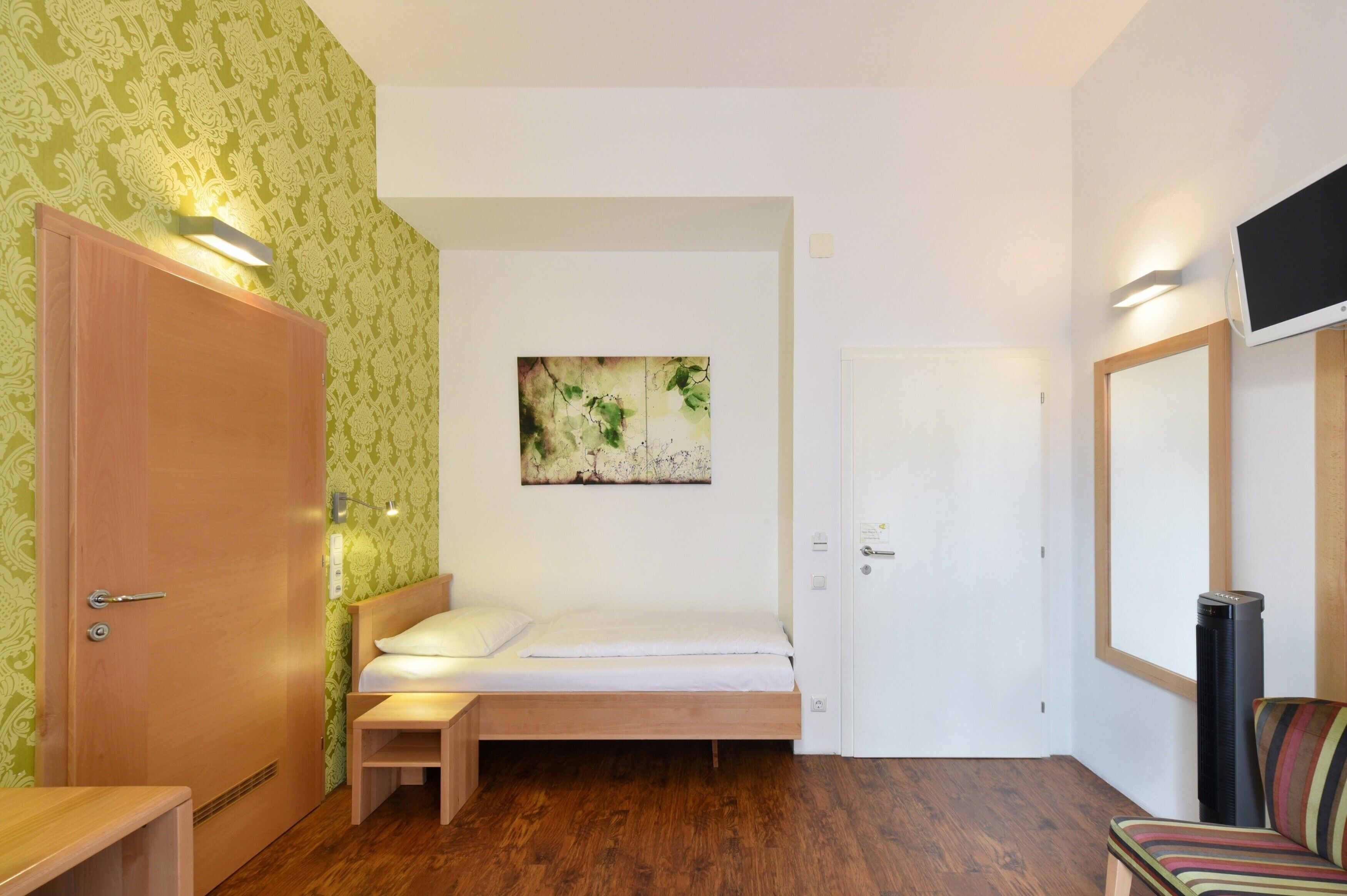 Ein Schlafzimmer mit einem hölzernen Bett, einem kleinen Nachttisch, einem an der Wand befestigten Fernseher und einer Tür mit Lichtschalter.