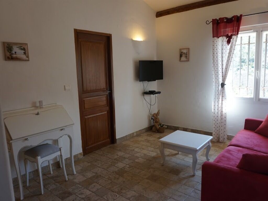 A room with a wooden door, a television mounted on the wall, a white table, a red sofa, and a window with curtains.