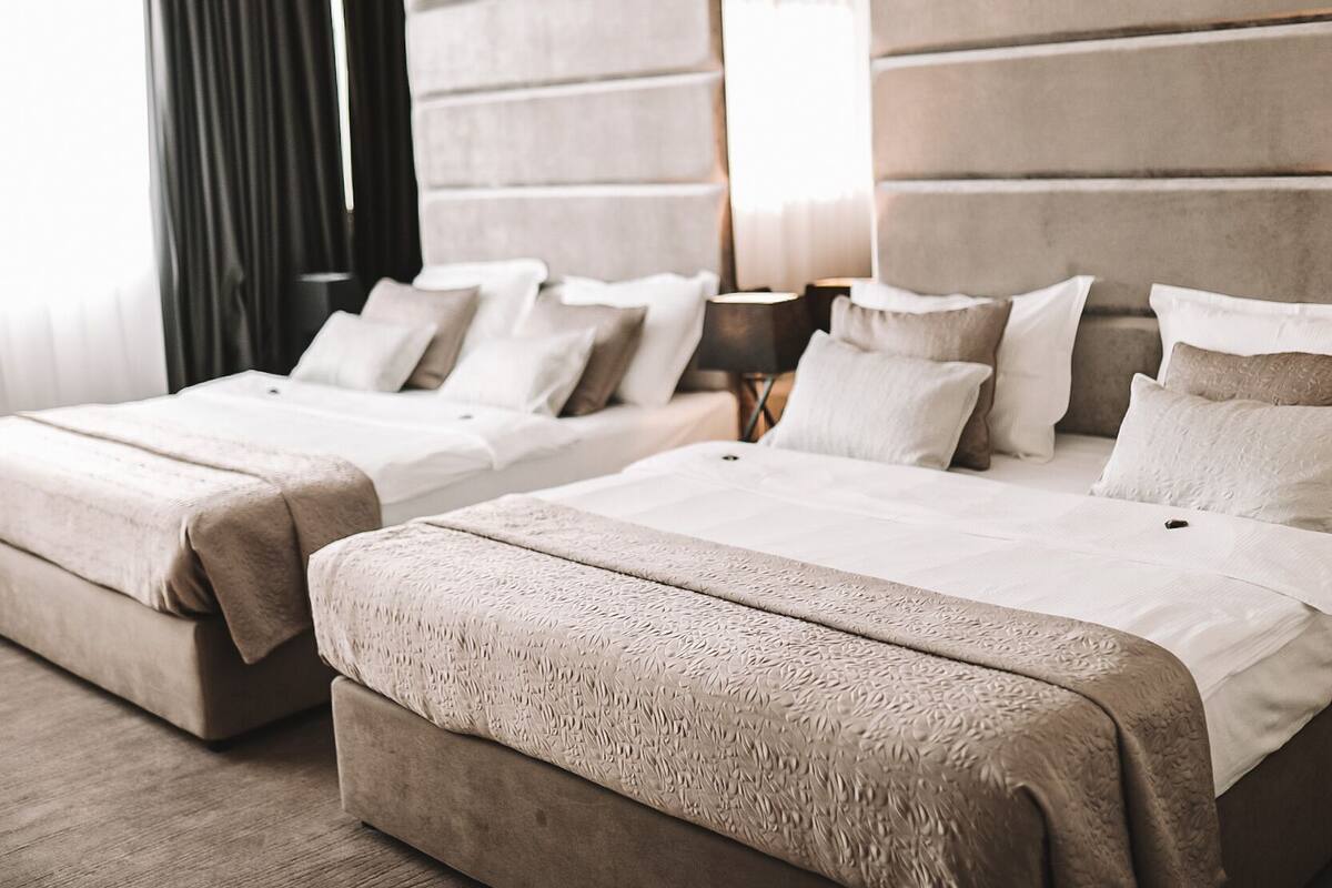 A hotel room with two beds, a large window with curtains, and a bedside table with a lamp.