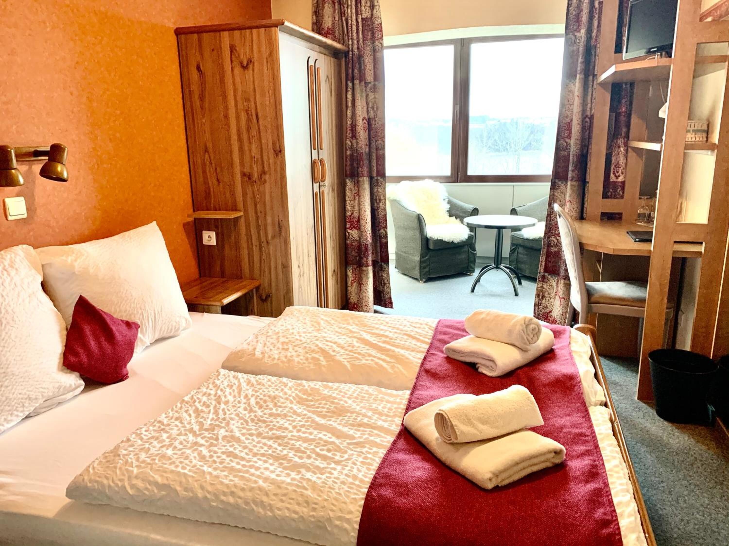 A hotel room with a large bed, a wooden wardrobe, a small table, and a view of the outdoors.