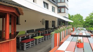 Un espacio al aire libre con mesas y bancos, un edificio de techo inclinado y un jardín con plantas.