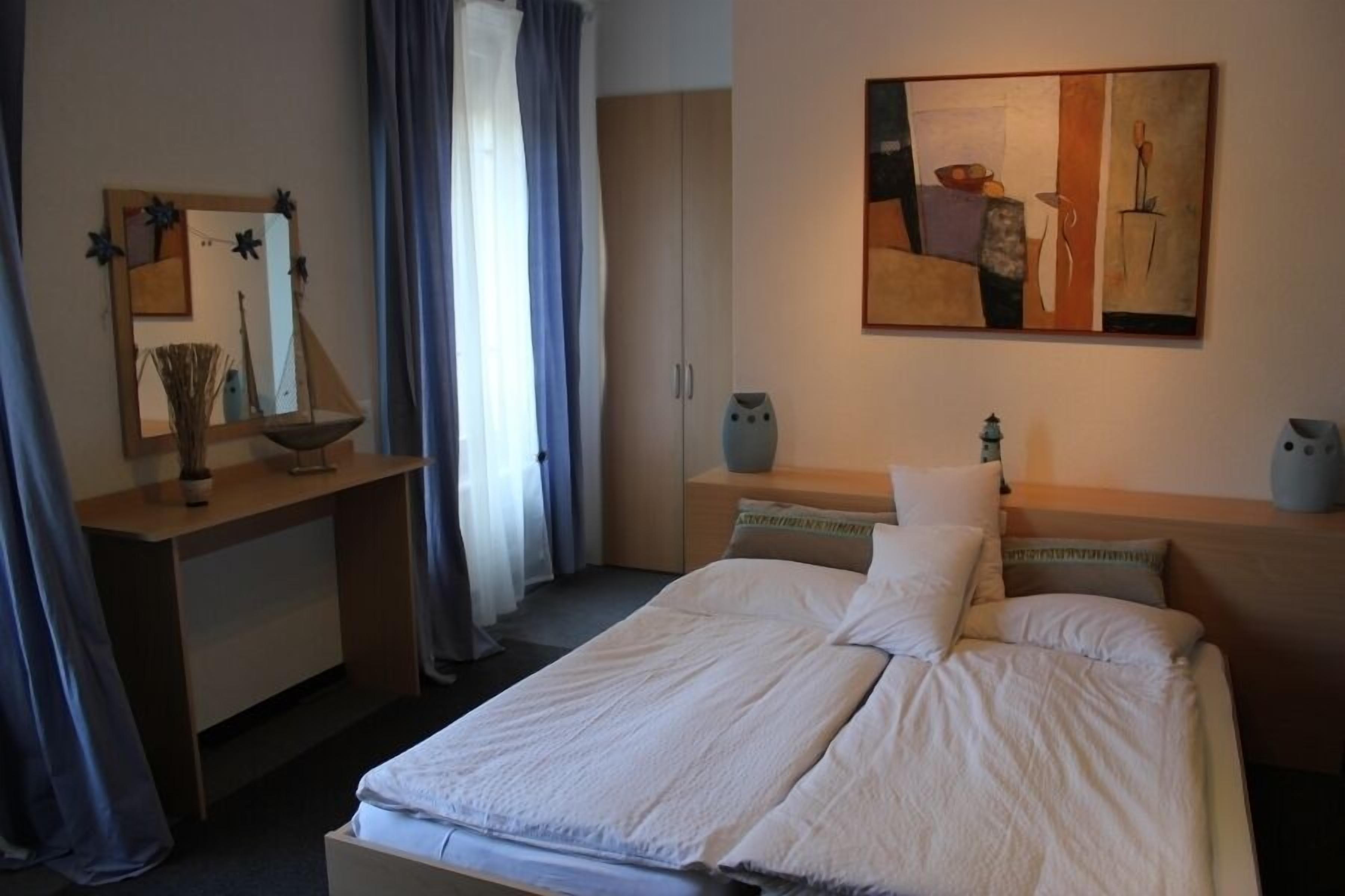 A hotel room with a bed, a desk, a chair, a wardrobe, and a painting on the wall.