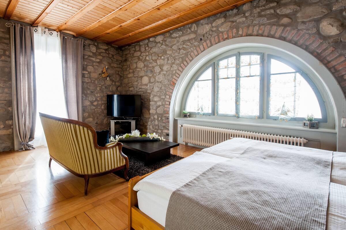 A bedroom with a stone wall, a large arched window, a bed, a chair, a TV, and a fireplace.