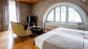 Une chambre avec un mur en pierre, une grande fenêtre en arc de cercle, un lit, une chaise, un téléviseur et une cheminée.