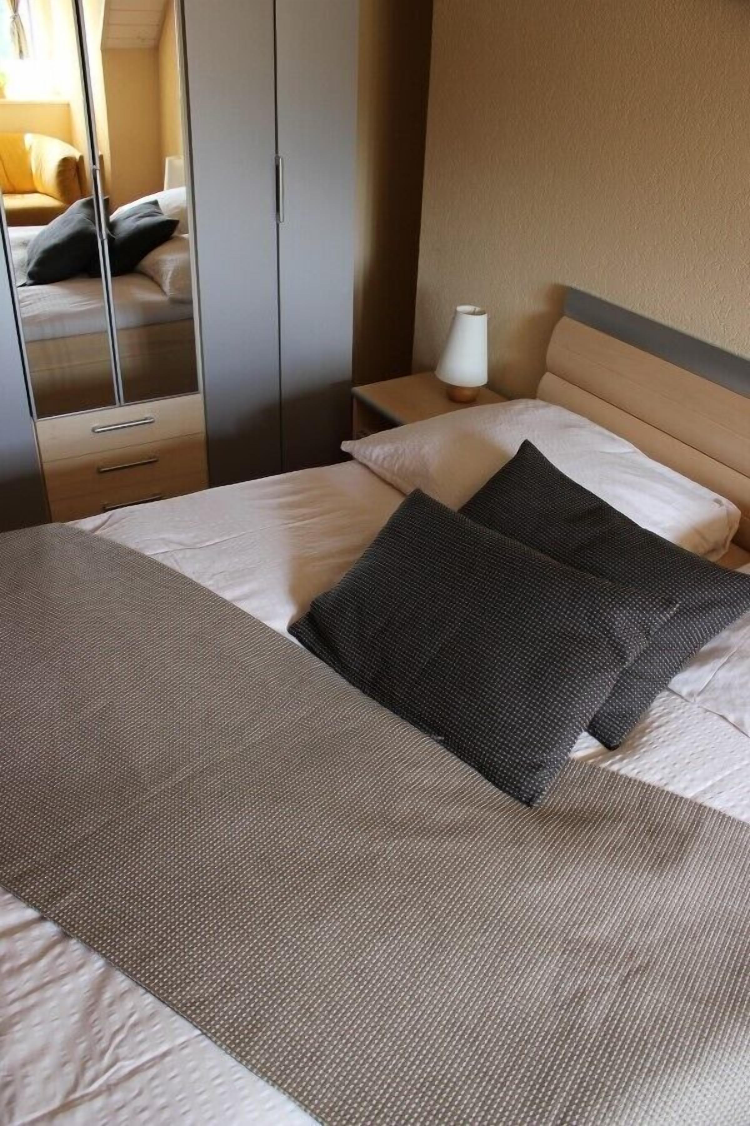 Un lit bien fait, avec une couette grise et blanche, une tête de lit en bois et une table de chevet sur laquelle se trouve une lampe.