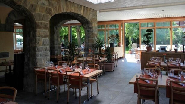 A restaurant with stone arches, large windows, and indoor plants. Tables are set with wine glasses and napkins. - Hotel Restaurant L'Acacia (Marly)