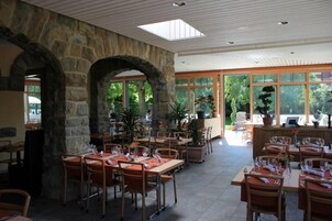 Un restaurante con arcos de piedra, amplios ventanales y plantas de interior. Las mesas están dispuestas con copas de vino y servilletas.