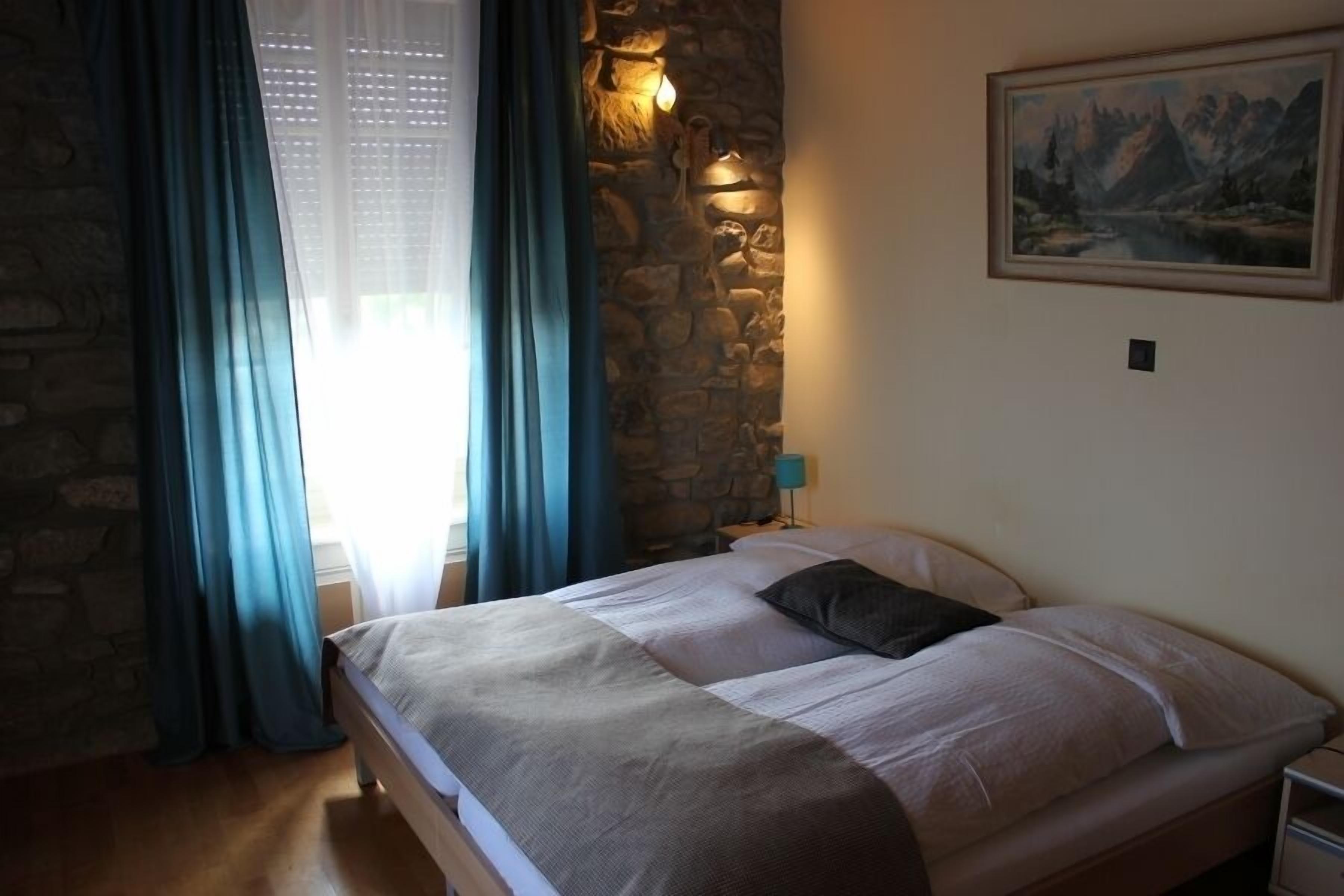 Un dormitorio con pared de piedra, una cama con edredón color beige y blanco, un cuadro en la pared y una mesita de noche con lámpara.
