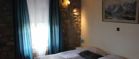 Un dormitorio con pared de piedra, una cama con edredón color beige y blanco, un cuadro en la pared y una mesita de noche con lámpara.