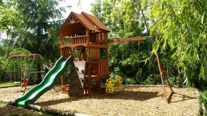 A wooden play structure with a slide and swings in a park. - Hotel Restaurant L'Acacia (Marly)