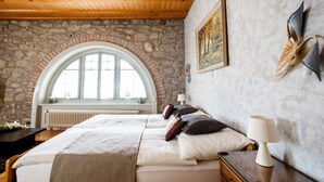 A bedroom with a stone wall, a large arched window, a bed with pillows, a bedside table, and a lamp. - Hotel Restaurant L'Acacia (Marly)