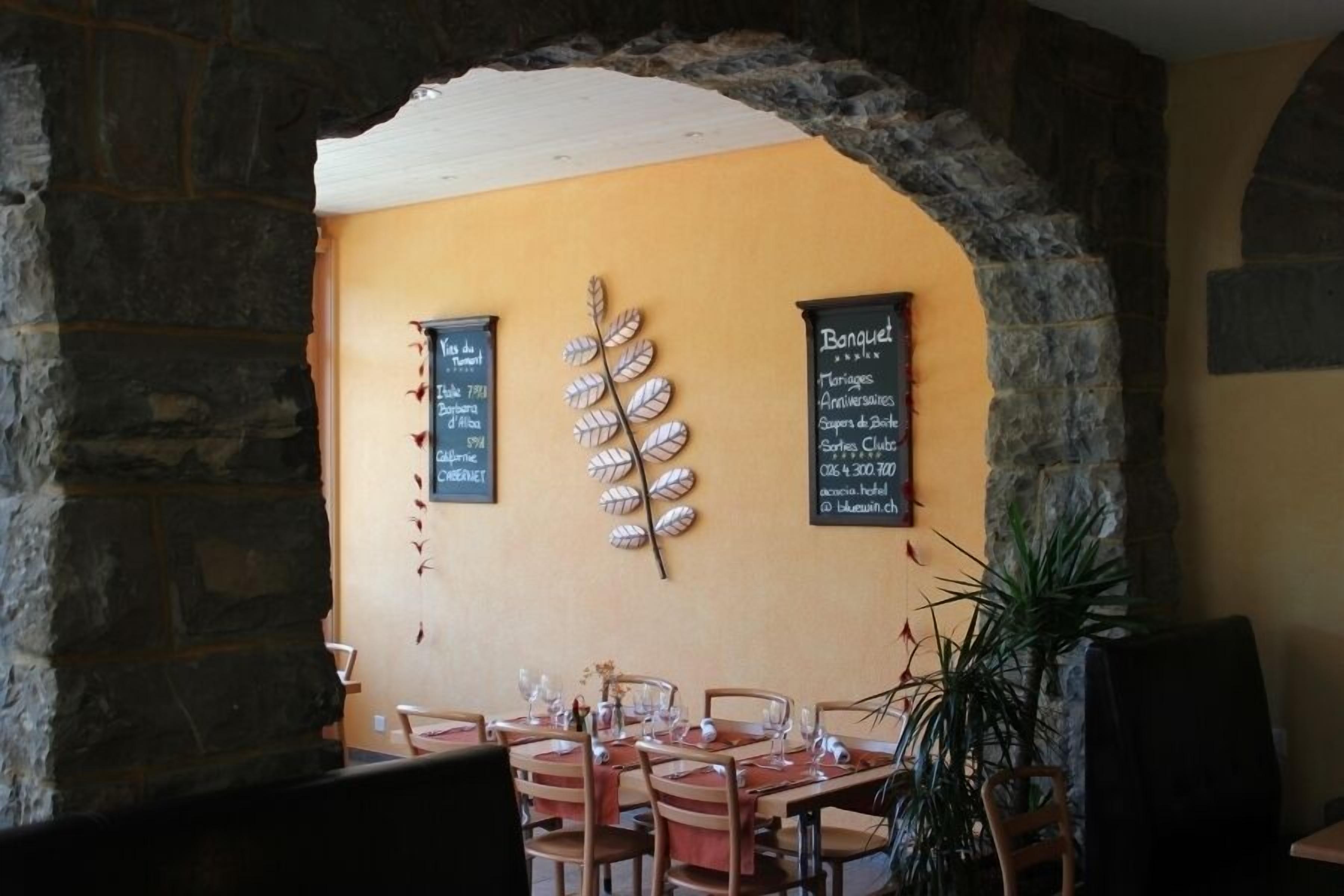 Ein Restaurantinneres mit Steinsäulen, Holztischen und -stühlen. Tafeln mit Speisekarten und eine dekorative Wandgestaltung mit Blättern sind zu sehen.