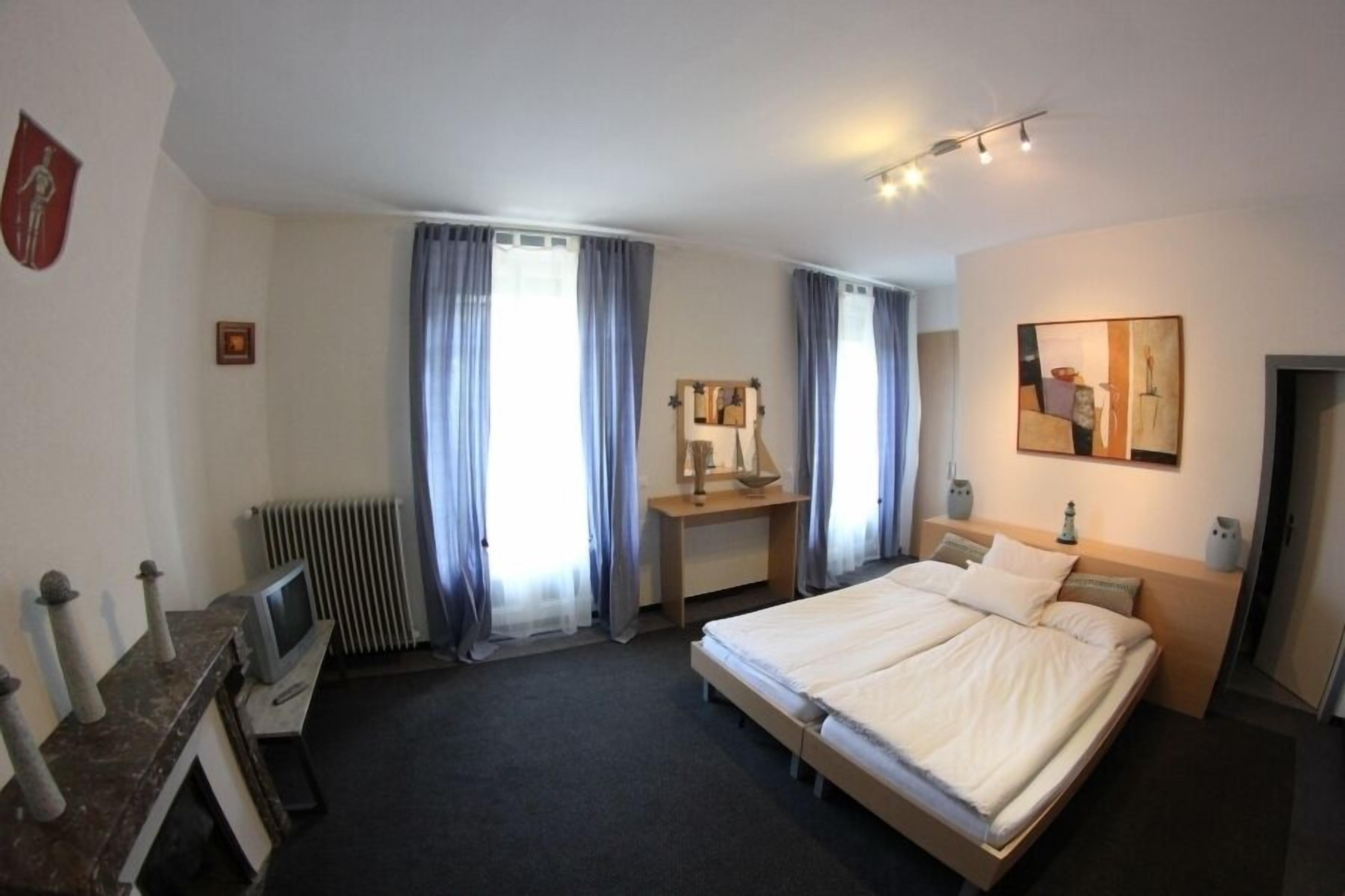 Una habitación de hotel con una cama, un escritorio con computadora, un radiador y cortinas.