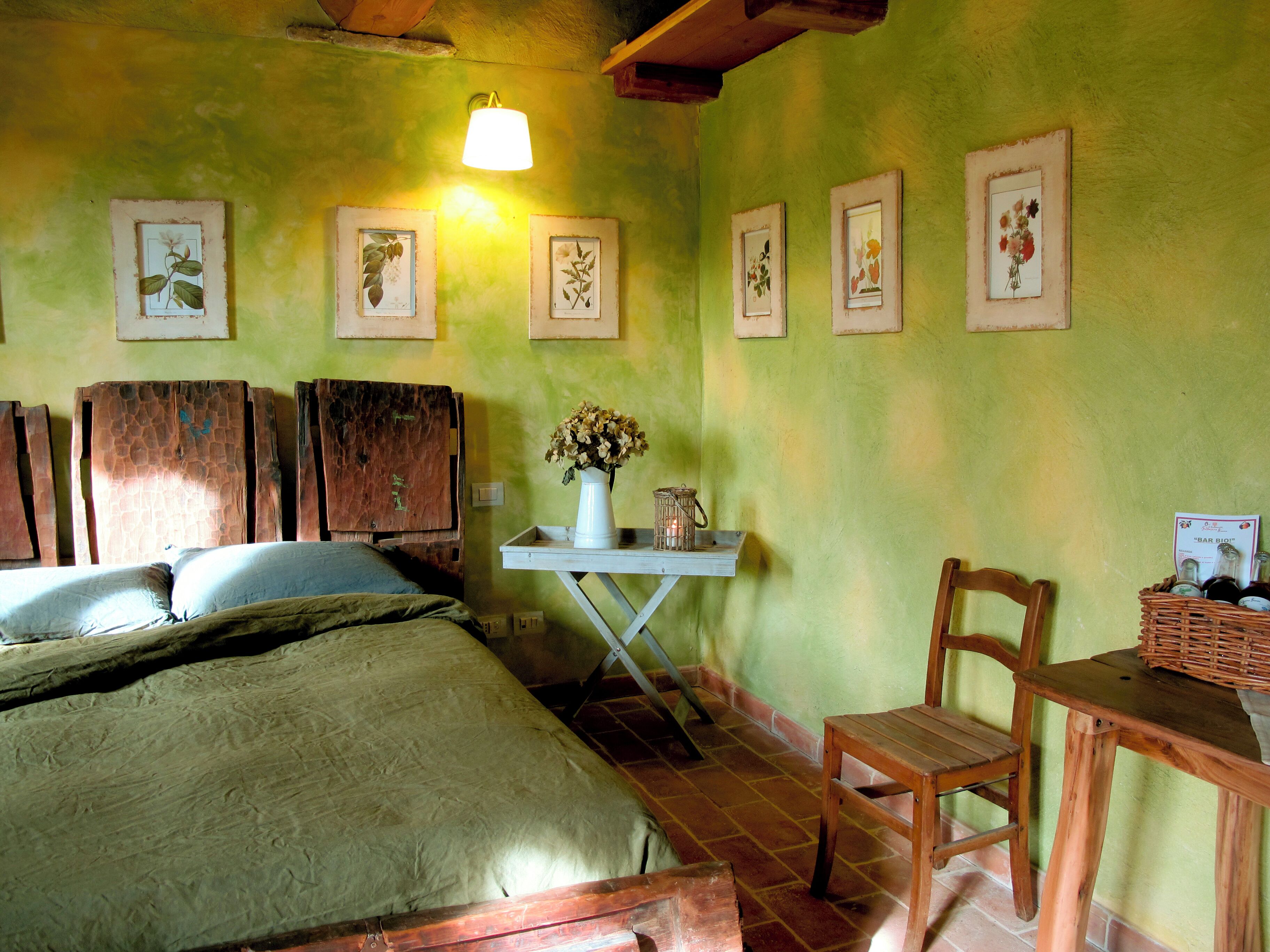 Une pièce comprenant un lit, une chaise en bois, une petite table avec un vase et une bougie, et des reproductions encadrées de motifs botaniques accrochées au mur.