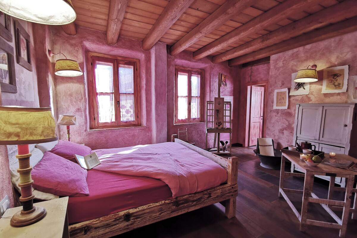 A rustic room with a wooden bed, a desk, and a chair. There are windows with curtains, a lamp, and a cabinet.