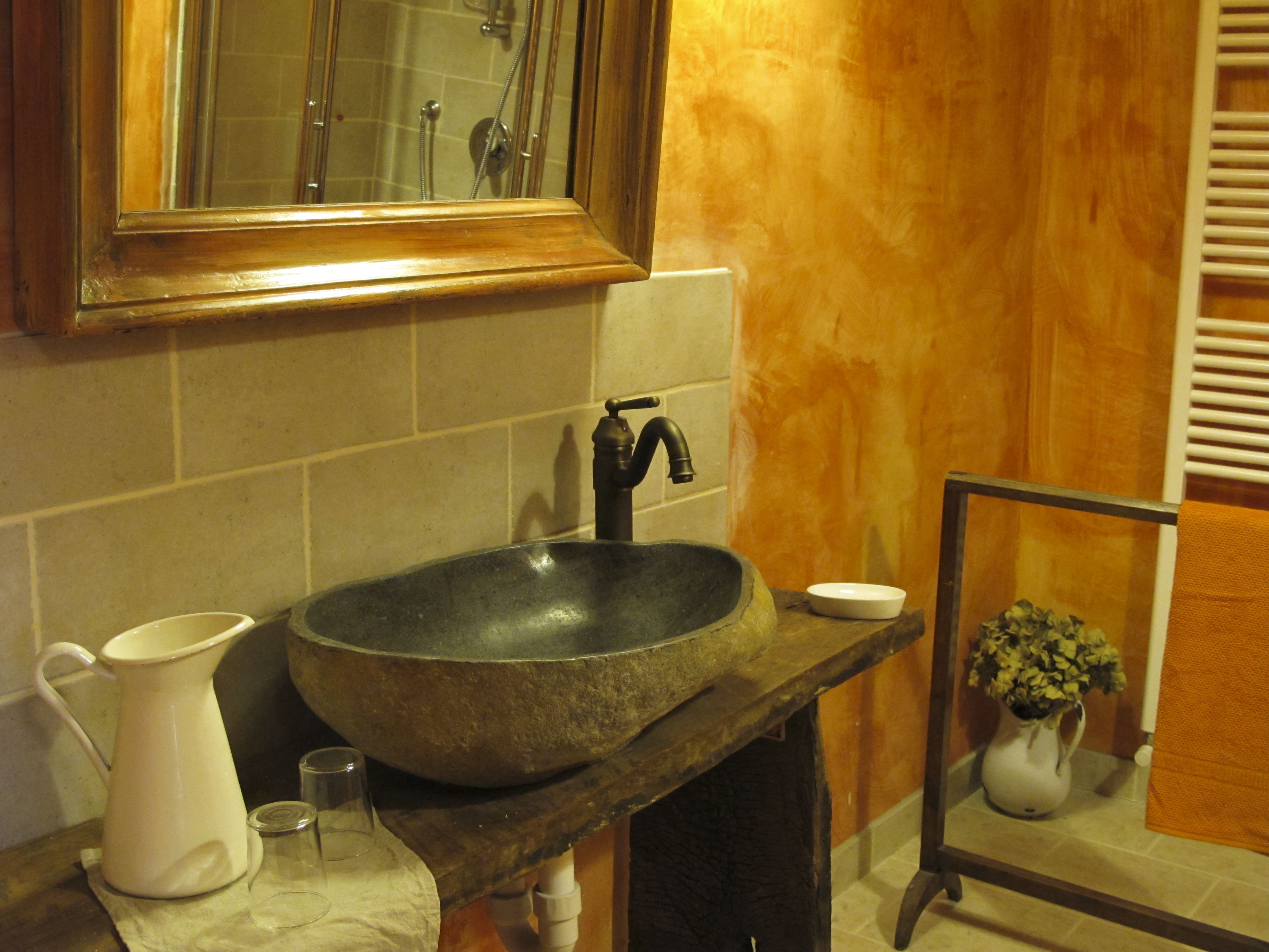 A bathroom with a stone sink, a mirror, a white pitcher, and a vase with flowers.