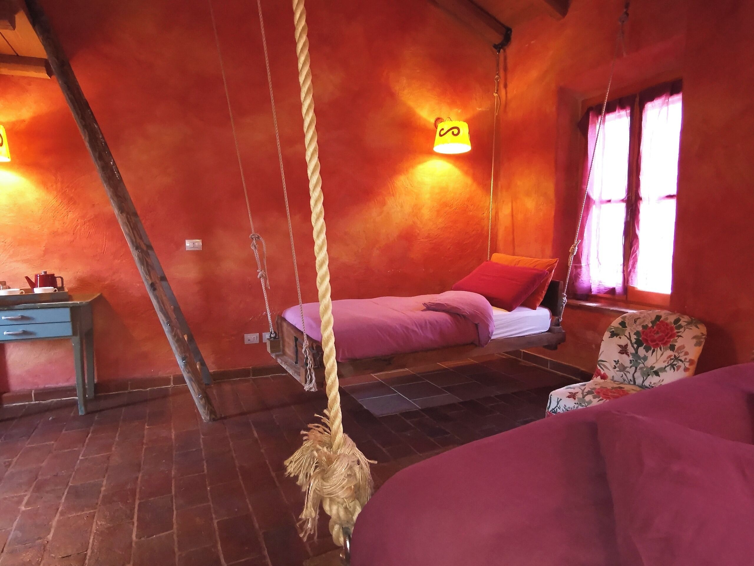 Ein rustikaler Raum mit einer Hängeschaukel, einem Bett mit violetter Bettwäsche, einem Holztisch mit einem roten Wasserkocher und einem blumigen Sessel.