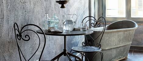 A rustic interior with a stone wall, a wooden ceiling, a table with chairs, a candle, and a vintage lamp.