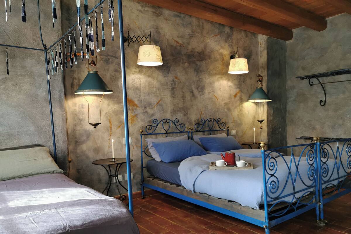 A rustic bedroom with a metal bed frame, two bedside tables, a wall-mounted lamp, and a hanging light fixture.
