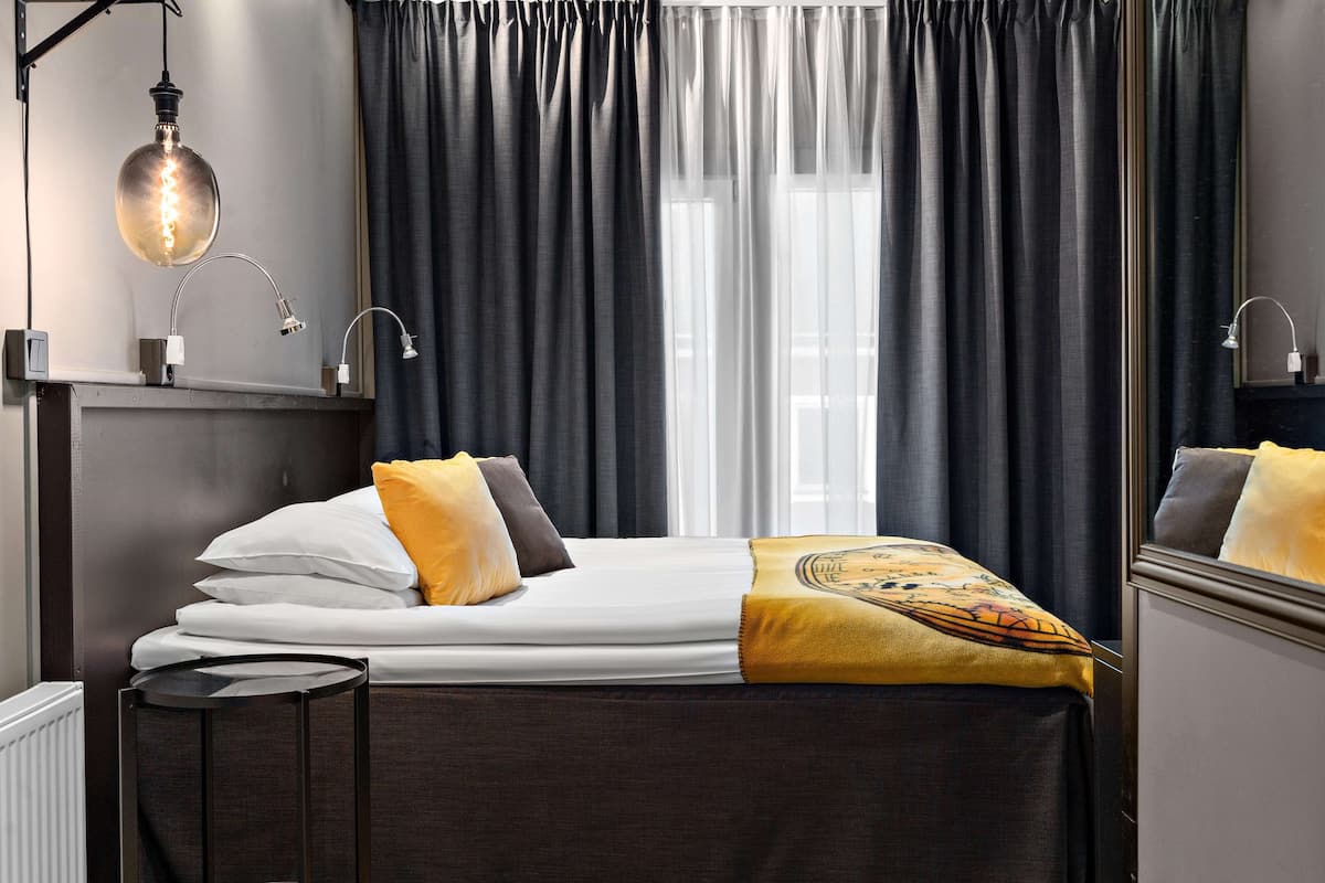 Ein ordentlich bezogenes Bett mit weißer und gelber Bettwäsche, ein Nachttisch mit Lampe und ein Fenster mit dunklen Vorhängen.