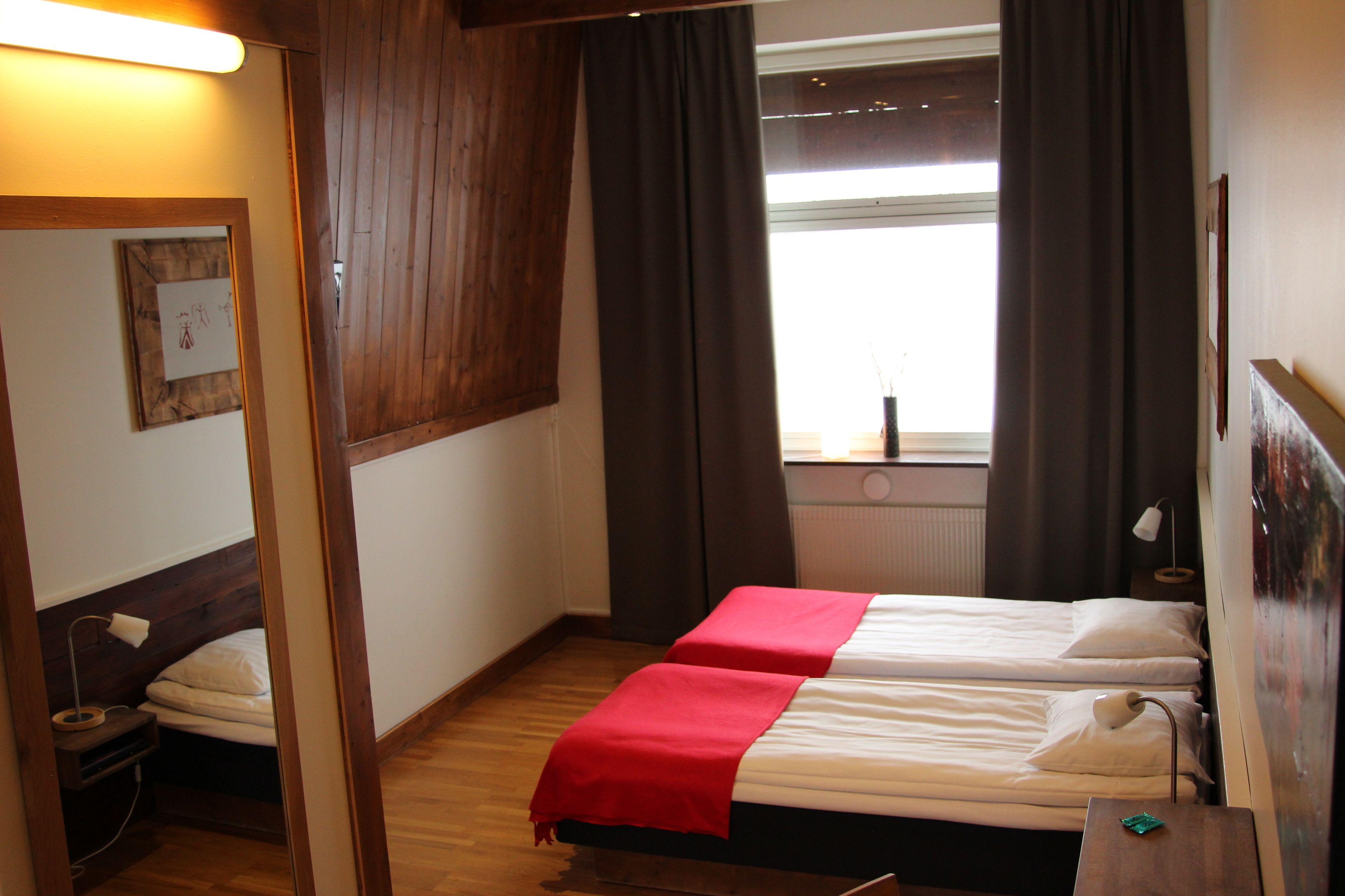A room with two single beds, wooden flooring, a window with curtains, a bedside table with a lamp, and a wall-mounted mirror.