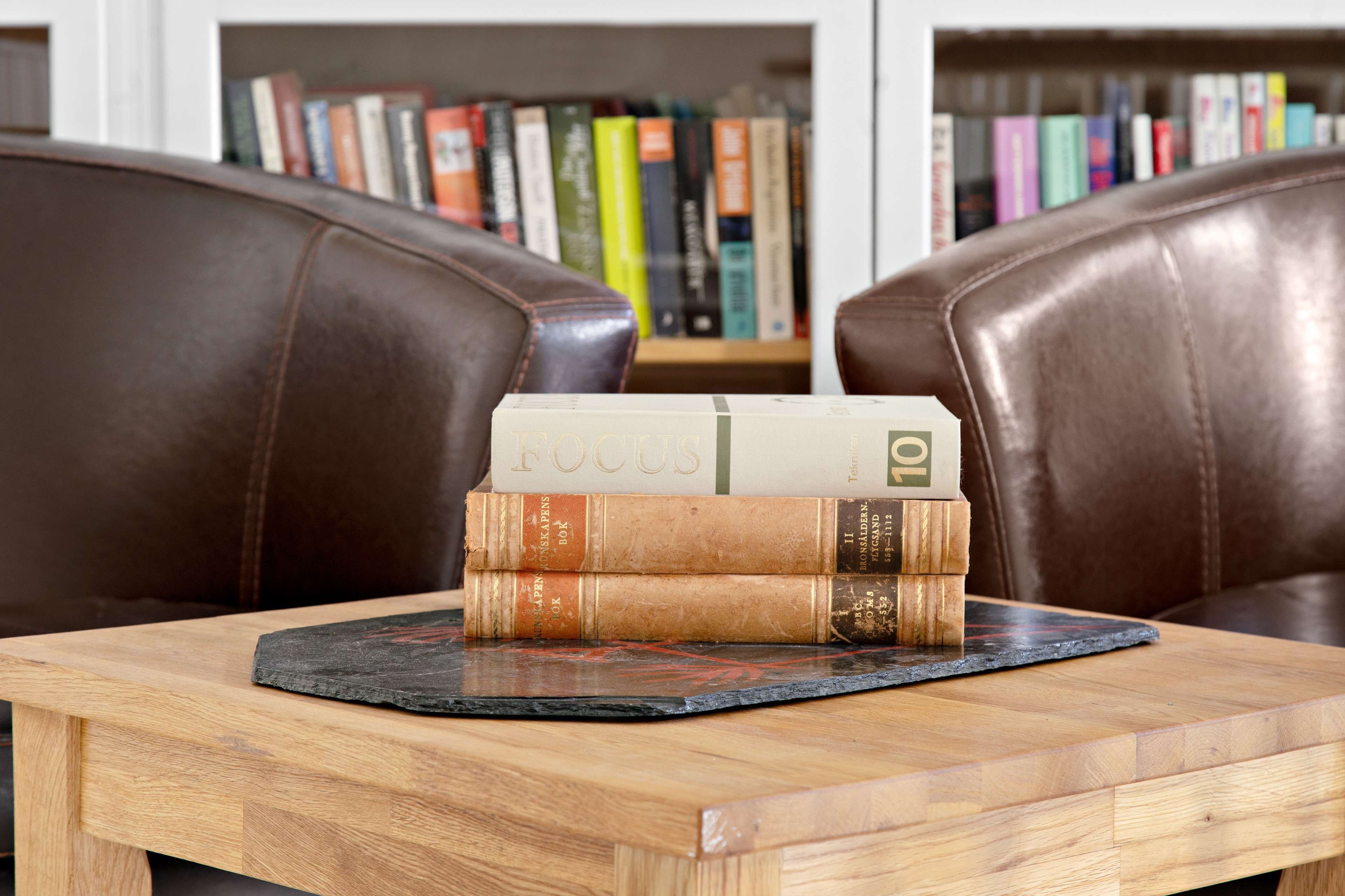 Una pila de libros sobre una mesa de madera con un mantel individual negro, frente a sillas de cuero marrón.