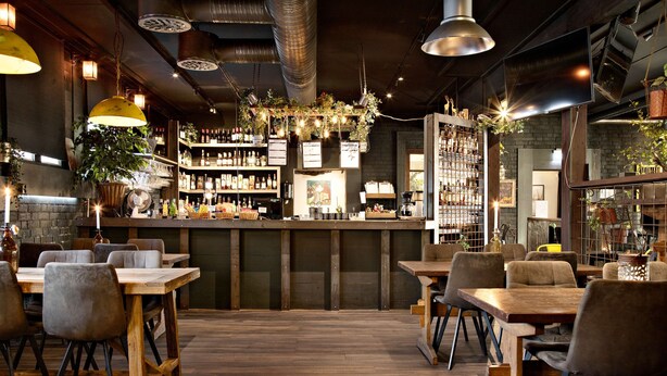 Um restaurante com mesas e cadeiras de madeira, um espaço de bar com prateleiras cheias de garrafas e plantas penduradas.