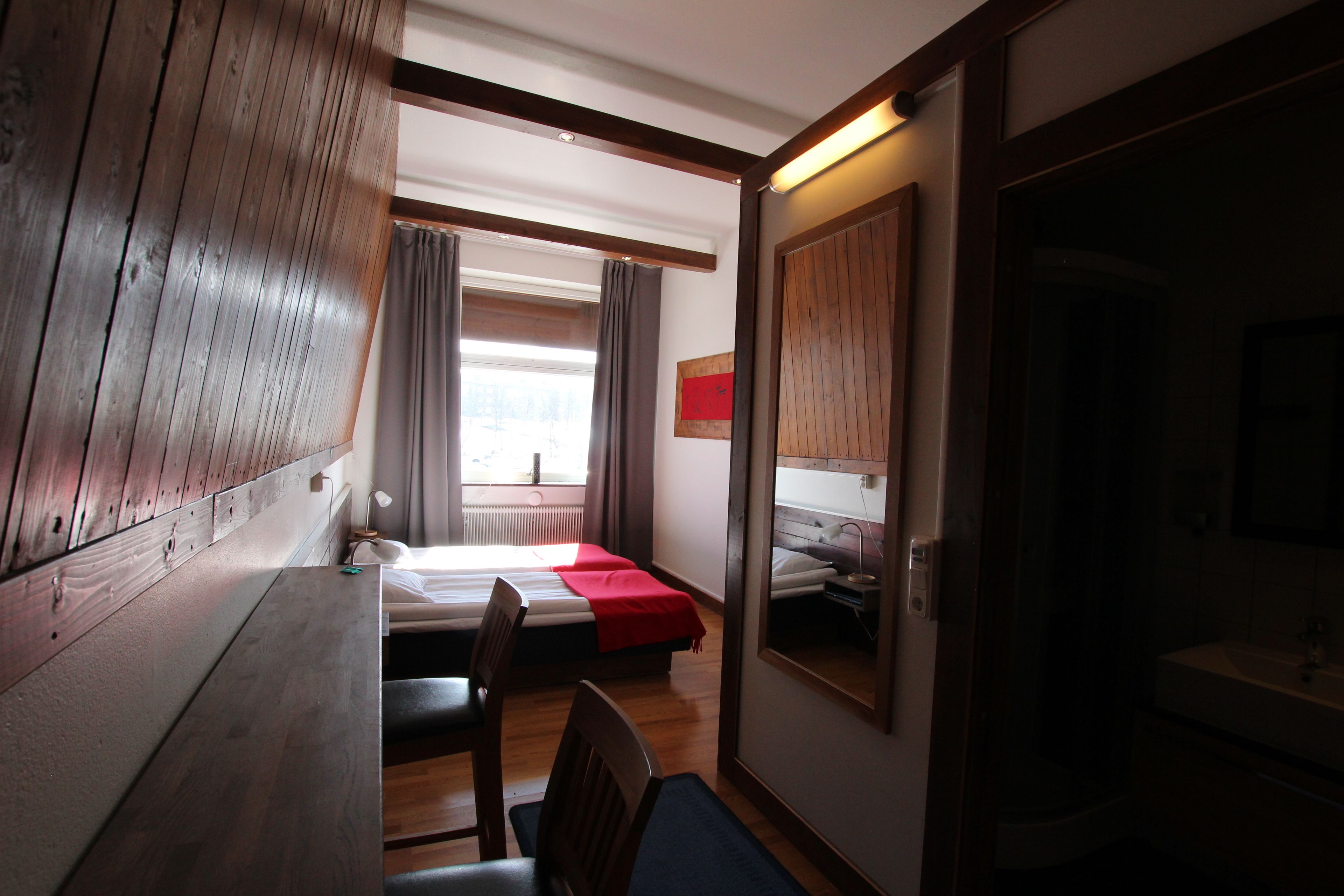 Una habitación con cabecera de madera, cama con manta roja, escritorio con silla, espejo y ventana con cortinas.