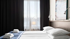 Quarto de hotel com duas camas, vista da cidade através de cortinas leves, um quadro na parede e uma mesa de cabeceira com abajur.