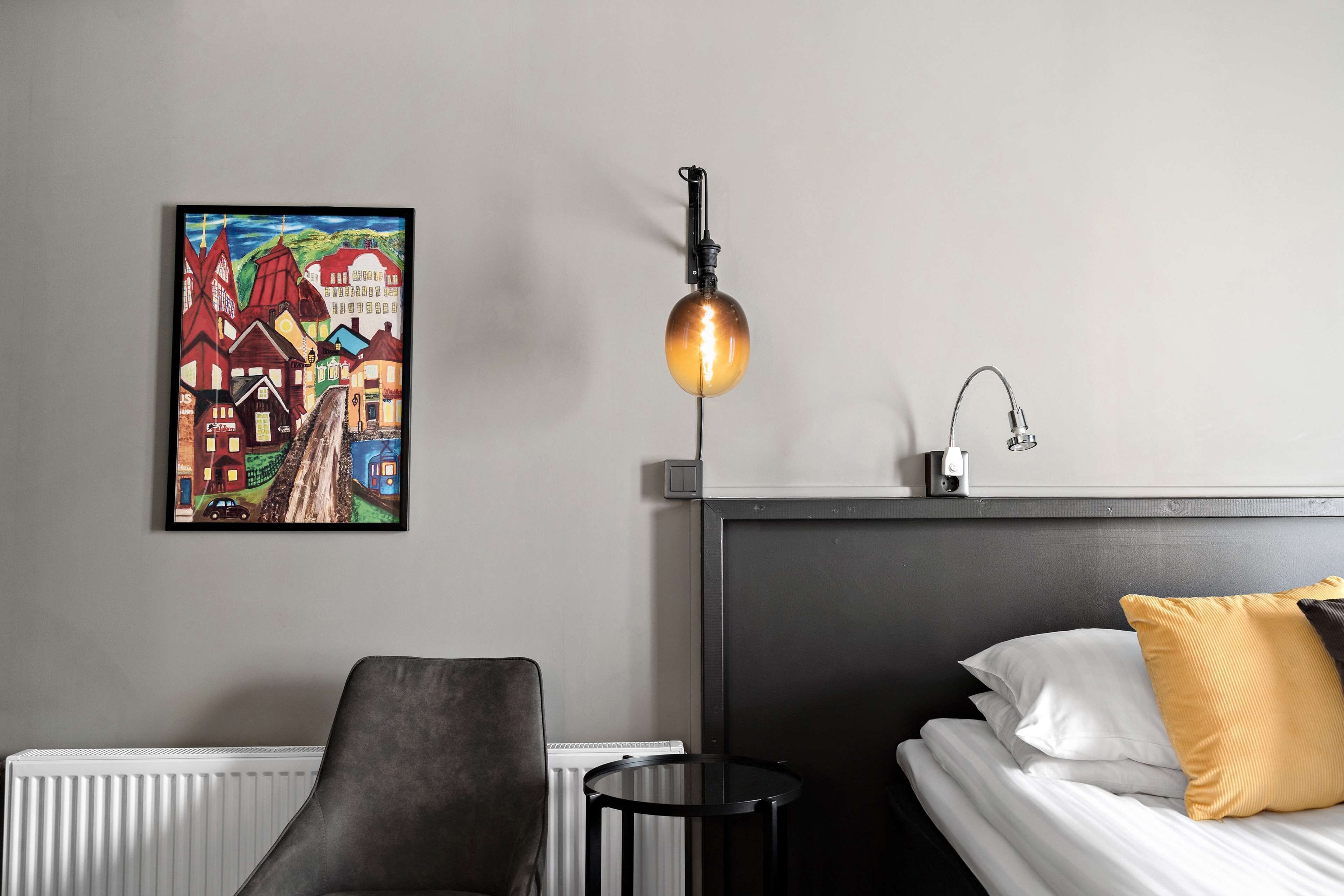 Hotellihuone, jossa on sänky, tuoli, pieni pöytä, patteri, seinään kiinnitetty valaisin ja värikäs taulu.