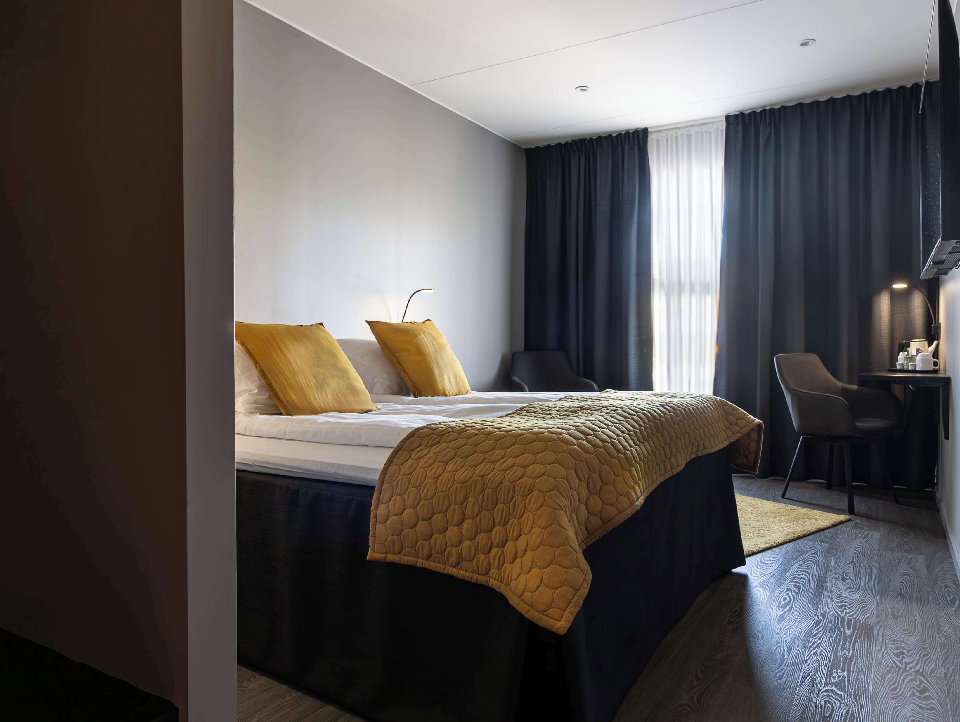 A modern hotel room with a bed, two pillows, a desk with a chair, and a window with curtains.