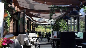 Um terraço ao ar livre com mesas e cadeiras, coberto por grandes guarda-sóis, e rodeado por vegetação.