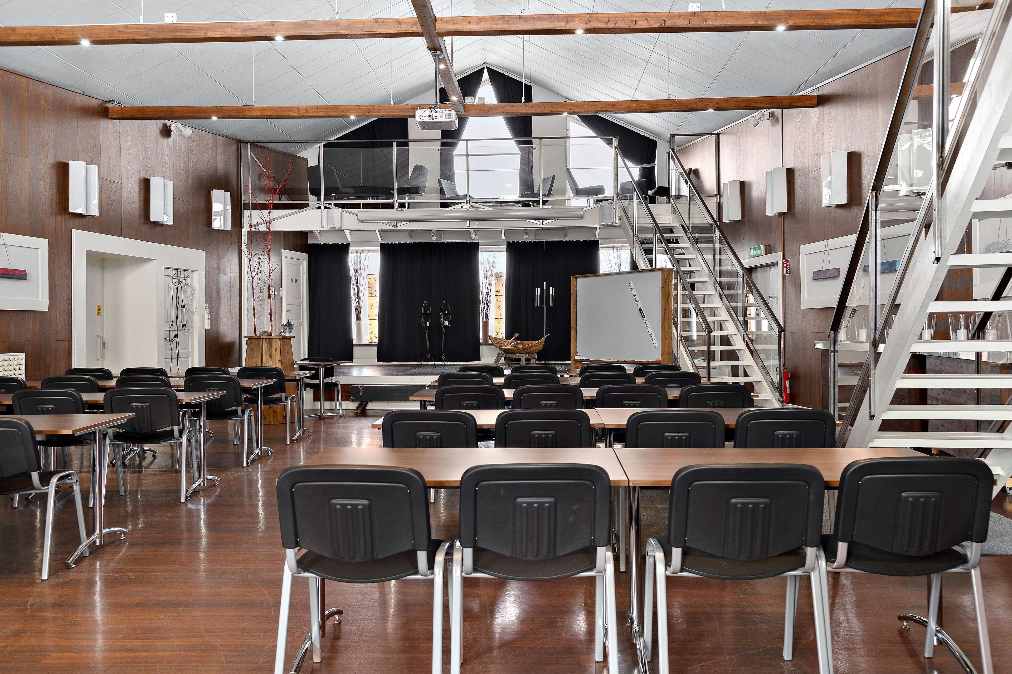 A spacious classroom with wooden floors, black chairs, and a stage with a whiteboard.