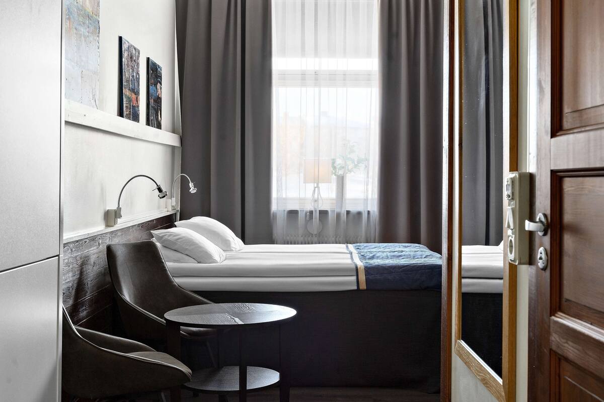 Ein kleines Hotelzimmer mit einem Einzelbett, einem Tisch, zwei Stühlen, einer Nachttischlampe und einem Fenster mit Vorhängen.