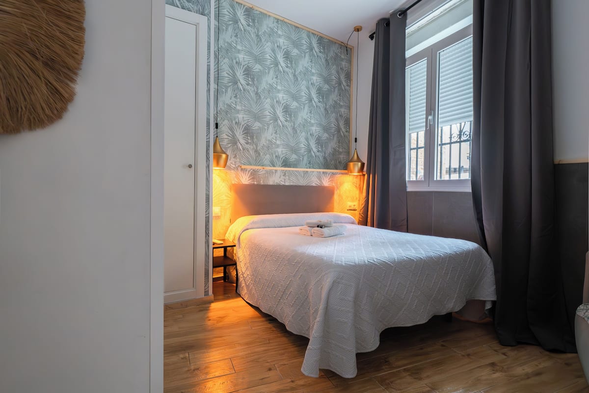 A bedroom with a bed, wooden floor, a wall with a leaf pattern, and a window with curtains.