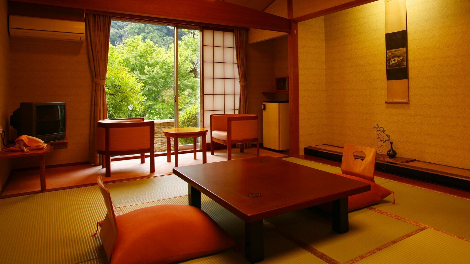 Una habitación de estilo japonés tradicional con suelo de tatami, una mesa baja, sillas, un televisor y vistas al exterior con vegetación.