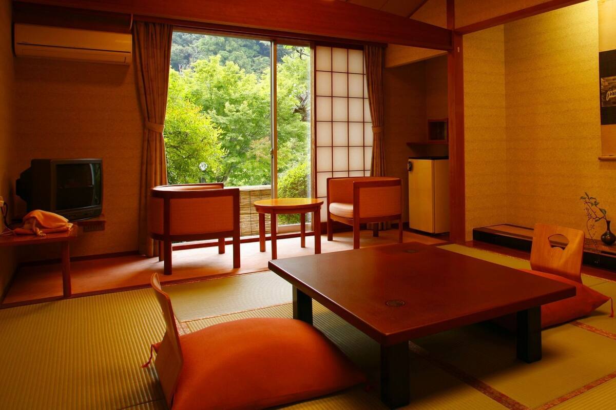 A traditional Japanese-style room with tatami flooring, a low table, chairs, a television, and a view of greenery outside.