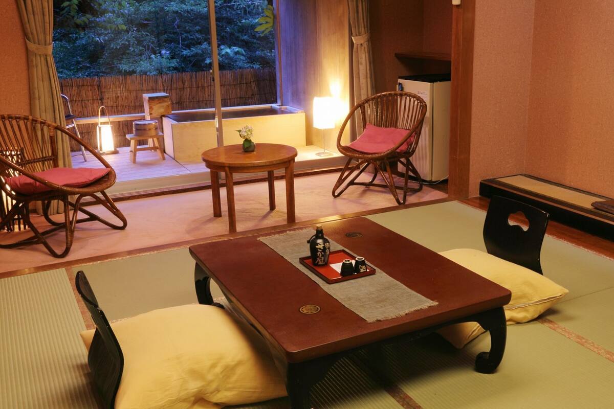 A traditional Japanese-style room with tatami flooring, a low table with a tray of sake, and two chairs with red cushions.