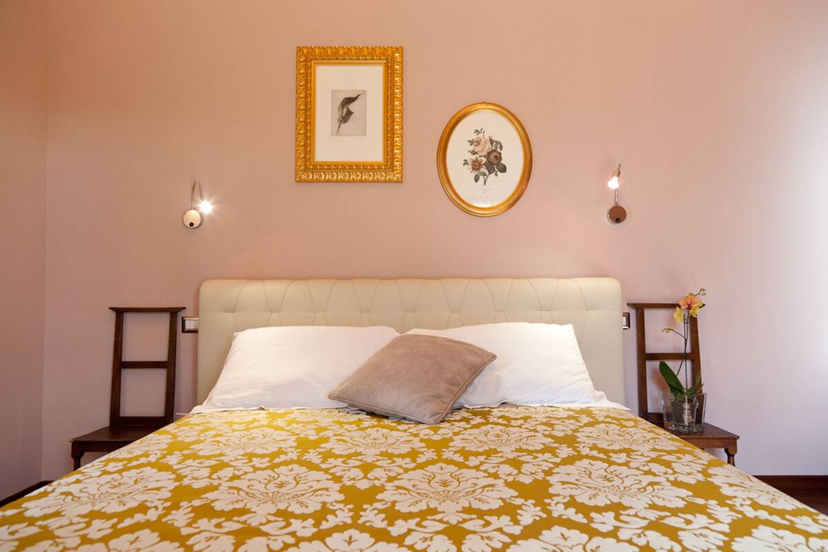A bedroom with a bed, two bedside tables, a nightstand, a wall-mounted lamp, and two framed pictures.