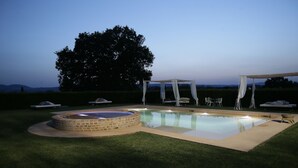Ein gut beleuchteter Außenpoolbereich mit Whirlpool, umgeben von einer Rasenfläche und einem großen Baum.