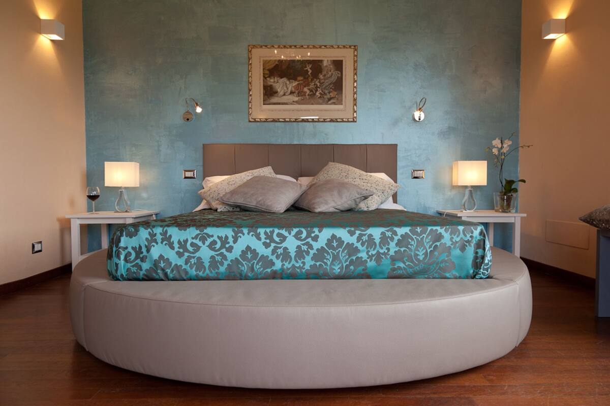 A modern bedroom with a round bed, two bedside tables, a nightstand, a framed artwork on the wall, and a vase with flowers on the nightstand.