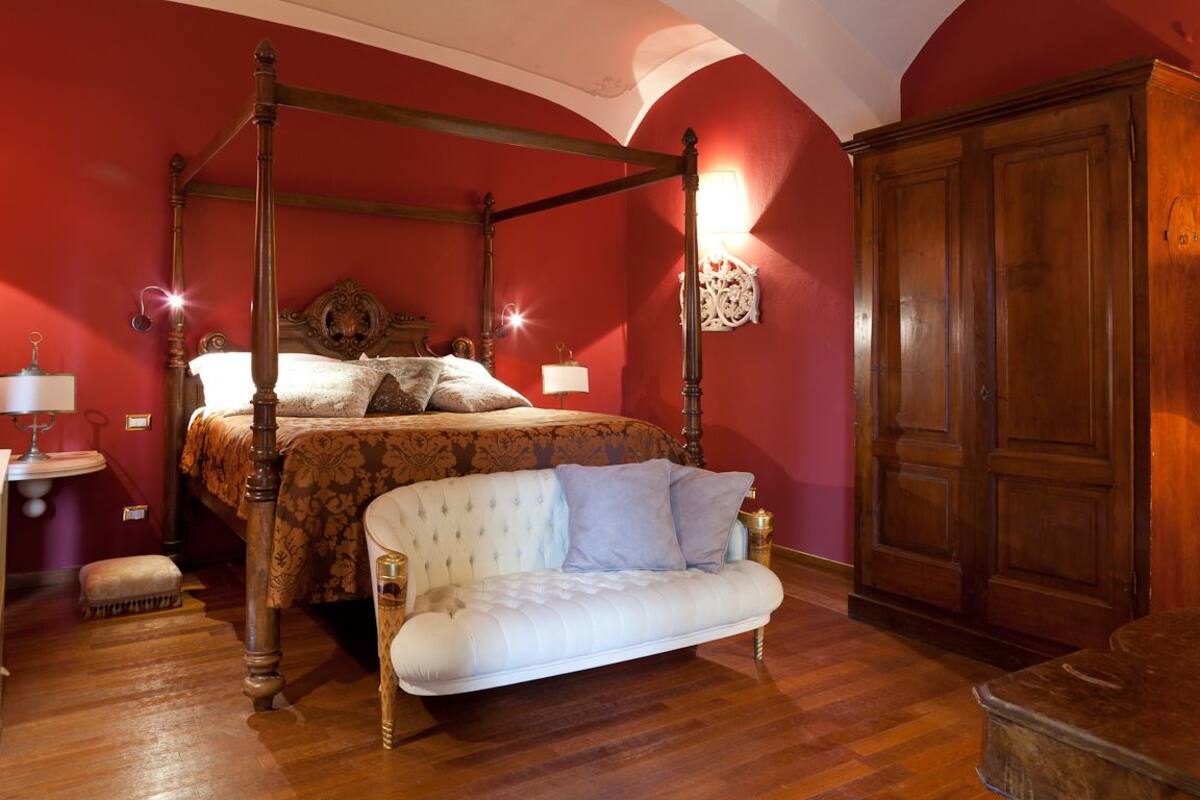 A four-poster bed with a canopy, a white upholstered sofa, and a wooden wardrobe in a room with red walls and wooden flooring.