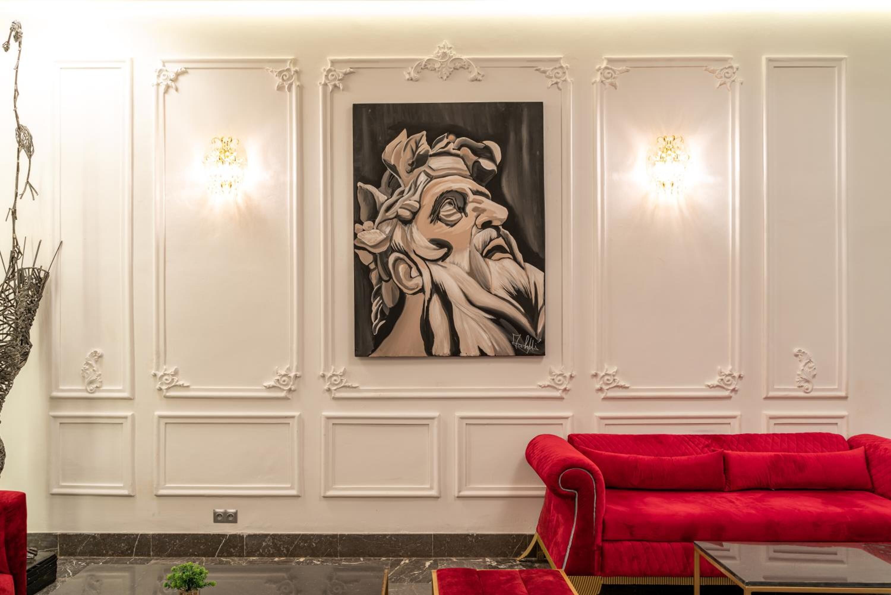 A room with a red sofa, a painting on the wall, and two wall sconces.