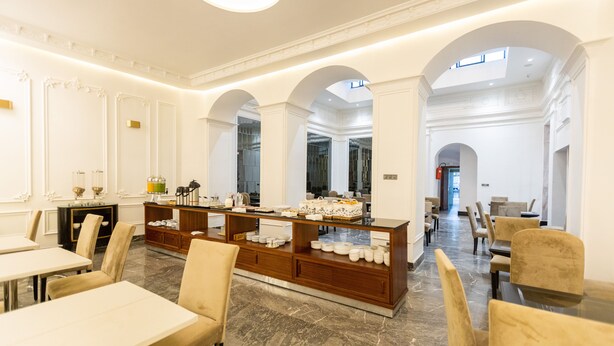 Amplia zona de comedor de hotel con suelo de mármol, paredes blancas y un bufé central con diversos platos.