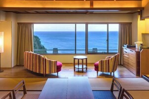 Una habitación con un ventanal grande con vista al océano, dos sillones a rayas, una mesita redonda y una mesa de comedor de madera con sillas.