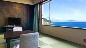 A room with a large window offering a view of the sea and hills, a flat-screen TV, a wooden table, and a traditional Japanese-style chair. - Yaizu Grand Hotel (Shizuoka)