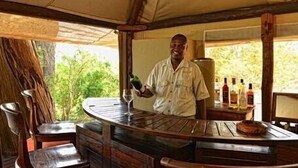 A wooden bar area with a person standing behind it, holding a bottle. There are chairs and a table with bottles on it.